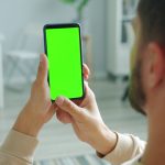 Pessoa segura um celular com a tela verde, em ambiente interno, para consulta ou uso de aplicativo.