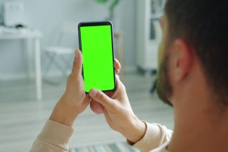 Pessoa segura um celular com a tela verde, em ambiente interno, para consulta ou uso de aplicativo.