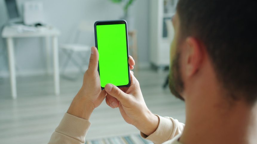Pessoa segura um celular com a tela verde, em ambiente interno, para consulta ou uso de aplicativo.