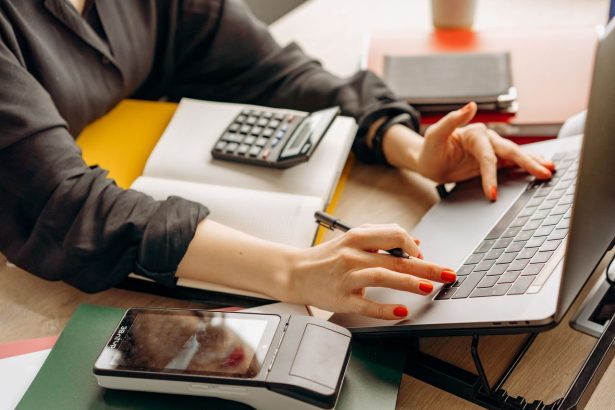 Pessoa usando um laptop ao lado de um terminal de pagamento e uma calculadora sobre a mesa.