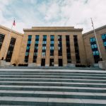 Exterior do prédio do Federal Reserve em Washington, com bandeiras dos Estados Unidos e escadaria sob céu nublado.