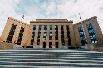 Exterior do prédio do Federal Reserve em Washington, com bandeiras dos Estados Unidos e escadaria sob céu nublado.