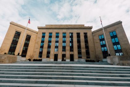Exterior do prédio do Federal Reserve em Washington, com bandeiras dos Estados Unidos e escadaria sob céu nublado.