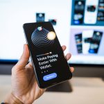 Mão segurando um smartphone com a interface de uma carteira digital aberta, diante de um monitor desfocado.