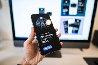 Mão segurando um smartphone com a interface de uma carteira digital aberta, diante de um monitor desfocado.