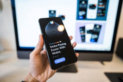 Mão segurando um smartphone com a interface de uma carteira digital aberta, diante de um monitor desfocado.